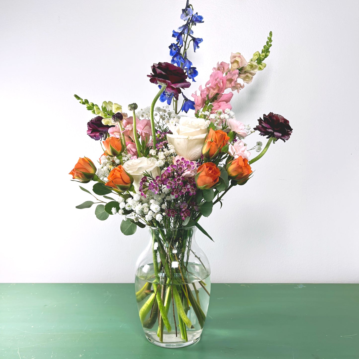 A vibrant floral arrangement featuring a variety of blooms including roses and other flowers in colors such as blue, pink, orange, white, and purple, placed in a clear glass vase.